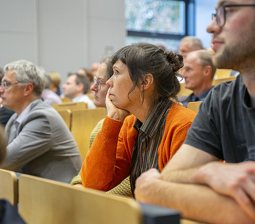 Die Sitzreihen sind mit Publikum besetzt, dass den Blick nach vorn richtet.