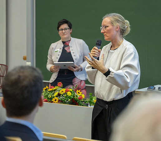Zwei Frauen stehen vor dem Publikum, eine spricht in ein Handmikro.