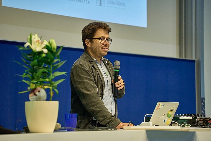 Prof. Weber mit Mikrofon in der Hand steht vor einem Pult im Hörsaal und hält eine Rede.