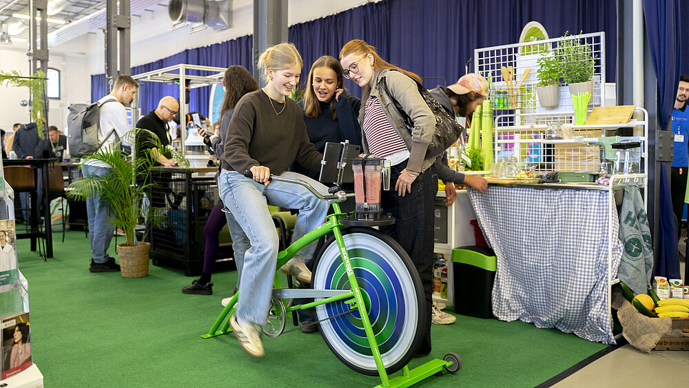 Eine Studieninteressierte radelt auf dem Smoothie-Bike, zwei Begleiterinnen stehen drumherum.