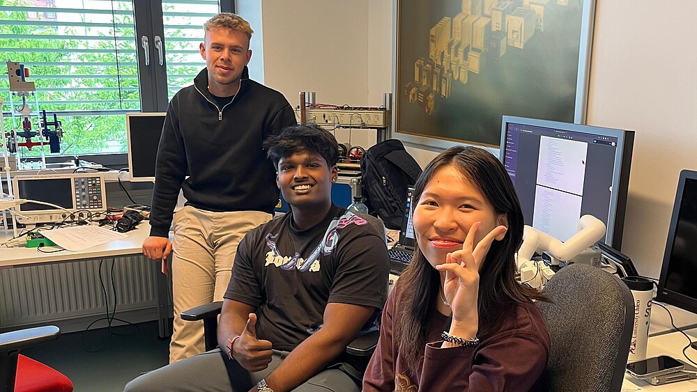 Drei ausländische Studierende sitzen in einem Büro und lächeln in die Kamera.