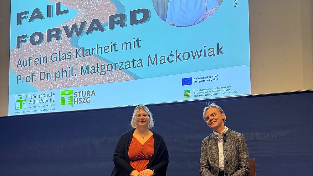 Aline Schulz und Prorektorin Mackowiak sitzen auf Hochstühlen vor einer blauen Hörsaaltafel. Darüber erscheint eine Beamerprojektion mit der Überschrift "Fail Forward".