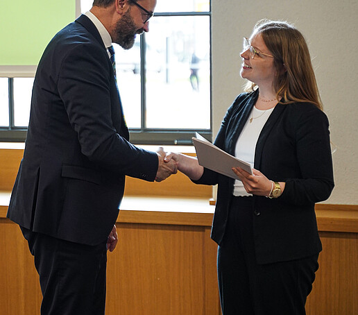 Ein Mann im Anzug schüttelt einer Frau in Businesskleidung die Hand. Beide lächeln sich an; die Frau hält eine Urkunde.